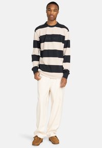 Gestreepte lange mouwen shirt in beige en zwart met een ronde hals; gecombineerd met een losse witte broek en bruine suède schoenen.