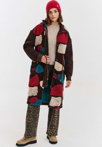 Žena v červenom pletenom čapici, multicolor patchwork fleecovej bunde, béžovom svetri, nohaviciach s leopardím vzorom a hnedých turistických topánkach stojí proti bielemu pozadiu.