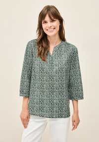 Grün gemusterte Bluse mit Dreiviertelärmel, die ein geometrisches Design in Weiß aufweist, locker geschnitten ist und einen Ausschnitt mit Schlitz hat.