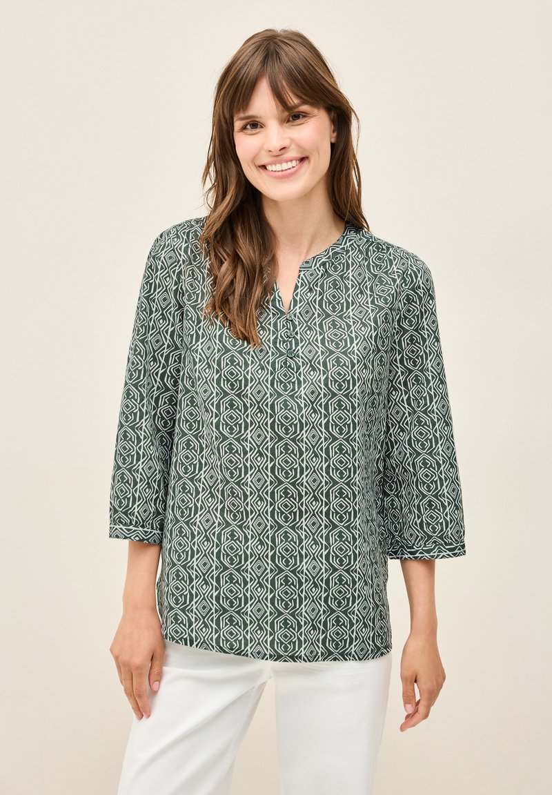 Grün gemusterte Bluse mit Dreiviertelärmel, die ein geometrisches Design in Weiß aufweist, locker geschnitten ist und einen Ausschnitt mit Schlitz hat.