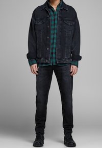 Chaqueta de mezclilla oscura con dos bolsillos en el pecho, sobre una camisa a cuadros verde y negra, combinada con jeans oscuros y botas de cordones negras.