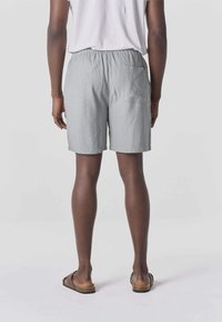 Persona con pantalones cortos de cintura elástica gris claro con un bolsillo trasero, camisa blanca y sandalias marrones, de pie mirando hacia atrás sobre un fondo gris.