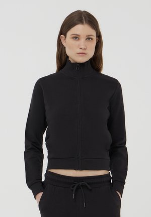 Giacca nera con zip, colletto alto e lunghezza corta, realizzata in tessuto liscio. Abbinata a pantaloni sportivi neri coordinati.