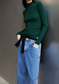 Maglione verde a coste con collo alto e maniche lunghe, abbinato a jeans in denim azzurro chiaro a vita alta, con una cintura nera di stoffa annodata in vita.