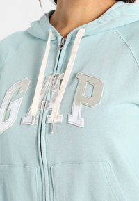 Hoodie zippé bleu clair en tissu doux. Il comprend une poche avant, des cordons blancs et des lettres "GAP" brodées brillantes sur la poitrine.