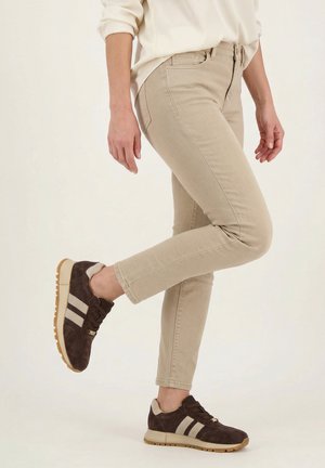 Person iført beige bukser og mørkebrune sneakers løfter det ene ben mod en ensartet lys baggrund.