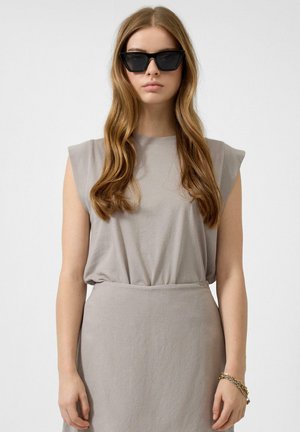 Vrouw met lang golvend haar, zwarte cat-eye zonnebril, mouwloos taupekleurig topje, bijpassende rok en een gouden kettingarmband om haar rechterpols.