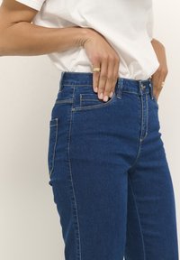 Jeans bleus taille haute en denim, au fit classique, avec cinq poches et des détails de couture ; texture lisse avec une légère élasticité.