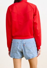 Röd sweatshirt med hög krage och ribbad nederkant, med chevron-sömmar på baksidan. Matchad med ljusblå denimshorts med bakfickor.