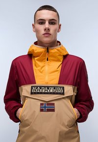 Farveblokket pulloverjakke med maroon og sennepsgule paneler, en lynlås krave, forlommer og et norsk flagg patch.