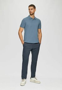 Mann steht mit den Händen in den Taschen, trägt ein blaues Polohemd, dunkle Hose und weiße Sneakers vor einem schlichten hellen Hintergrund.