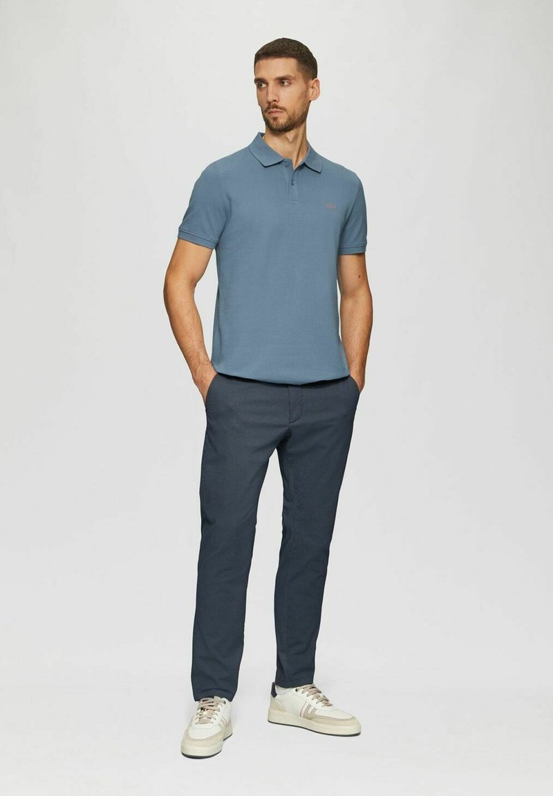 Mann steht mit den Händen in den Taschen, trägt ein blaues Polohemd, dunkle Hose und weiße Sneakers vor einem schlichten hellen Hintergrund.