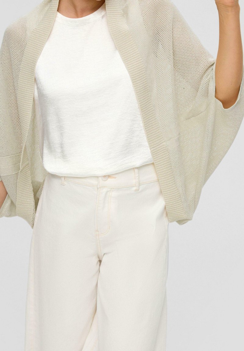 Personne portant un cardigan en maille beige ample par-dessus un t-shirt blanc rentré dans un pantalon taille haute écru, sur fond uni.