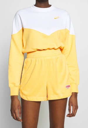 Persoon draagt een gele en witte Nike-sweatshirt met bijpassende gele shorts met een roze Nike-logo op het linkerbeen.