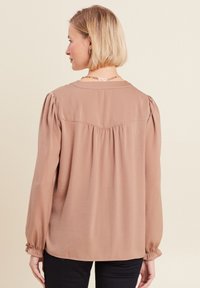 Blusa a maniche lunghe in morbido tessuto beige, con scollatura rotonda, spalle arricciate e polsini elasticizzati. Il retro presenta dettagli di pieghe sottili.