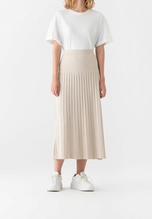Woman wearing a white short-sleeve top, beige high-waisted pleated midi skirt, and white sneakers standing against a plain background.