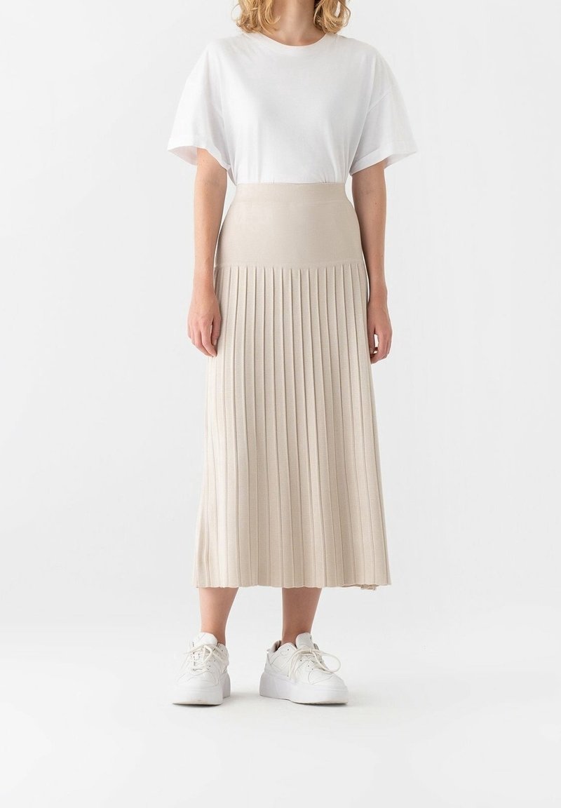 Femme portant un haut blanc à manches courtes, une jupe midi plissée beige taille haute et des baskets blanches, debout devant un fond uni.