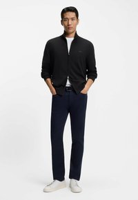 Mężczyzna stojący z rękami w kieszeniach, ubrany w czarny sweter na zamek, białą koszulę, granatowe spodnie, czarny pasek i białe trampki na białym tle.