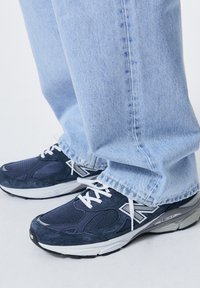 Zapatillas New Balance en color marino, con parte superior de malla y ante, cordones blancos y suela gris, combinadas con jeans de mezclilla azul claro.