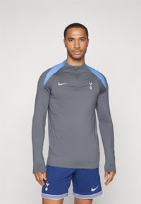 Langärmliges graues Sportshirt mit blauen Akzenten und einem Reißverschlusskragen, versehen mit einem Vereinslogo auf der Brust, getragen mit blauen Shorts.