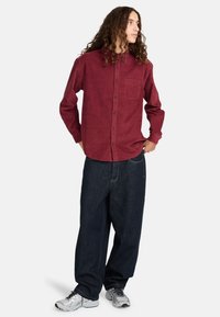 Burgunderfarbendes Cordhemd mit Button-Down-Kragen, Brusttasche, kombiniert mit locker sitzenden dunklen Denim-Hosen und grauen Sportschuhen.