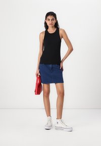 Zwarte mouwloze top gecombineerd met een blauwe spijkerrok en een rode handtas. Model draagt witte hoge sneakers. Eenvoudige, casual outfit tegen een witte achtergrond.