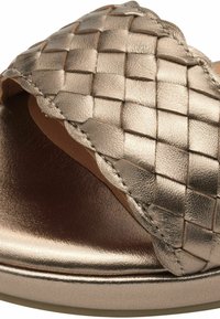 Sandaña metálica dorada con un diseño de parte superior trenzada, suela plana y una textura suave. Los acentos incluyen costuras a lo largo de los bordes.