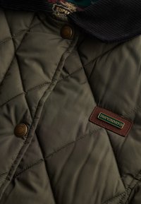 Gequilted olijfgroene jas met een corduroy kraag, drukknoopsluiting en een bruine label met "SUPERDRY" in gouden letters.