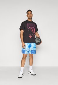 Svart grafisk t-skjorte med lilla print, blå mønstrede shortser, hvite sokker og hvite joggesko. Holder en svart basketball.