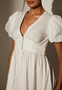 Weißes Kleid aus leichtem Stoff mit gepufften kurzen Ärmeln, einem tiefen V-Ausschnitt und einer durchgehenden Knopfleiste mit gerafften Details an der Taille.