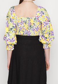 Blouse à imprimé floral avec des manches bouffantes en jaune, violet et vert, dotée d'un dos carré et de poignets élastiques, associée à une jupe noire.