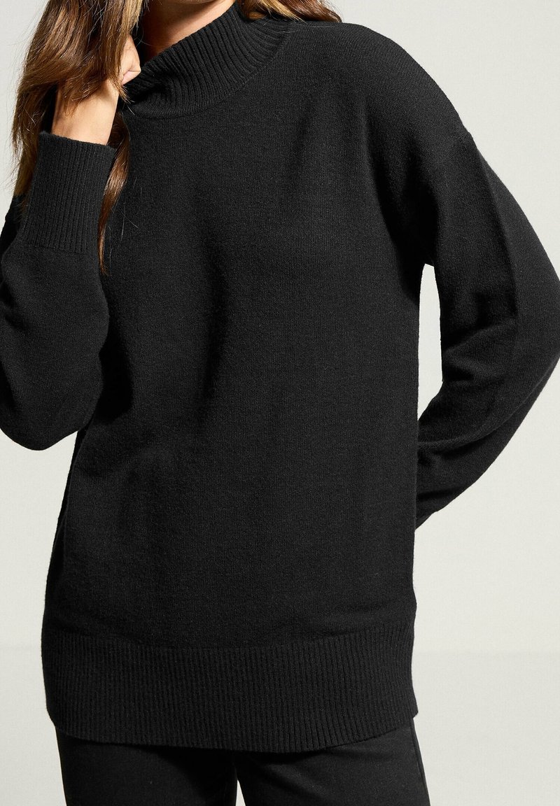 Personne portant un pull à col roulé côtelé noir à manches longues, main touchant le visage, sur un fond clair uni.