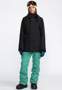 Veste imperméable noire, pantalon bleu sarcelle et bottes noires. Comprend un col haut, des poches spacieuses et des gants texturés. Lunettes de ski sur la tête.