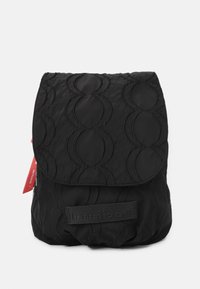 Mochila negra de nailon con un patrón de ondas acolchadas, una solapa plegable y un parche con el logotipo en la parte frontal. Incluye una etiqueta roja en contraste.