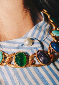 Collana in oro con pietre ovali turchesi, verdi e blu. È visibile una camicia a righe blu e bianche con un grande bottone di perla.
