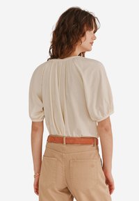 Ecrufarbenes kurzärmliges Blusenshirt mit gerafftem Rückendetail und elastischen Bündchen, kombiniert mit beigen hoch taillierten Hosen und einem hellbraunen Gürtel.
