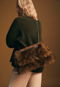Donna bionda che indossa un maglione verde scuro e pantaloni chiari, con una grande borsa in pelliccia marrone e catena dorata a tracolla.