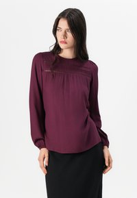 Bordeauxblus med långa puffärmar, rund halslinning och draperad detalj vid bröstet. Slät tygkvalitet, något lös passform, minimala accenter.