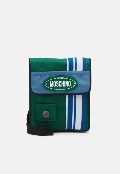 Sac bandoulière en tissu vert et bleu avec un rabat à l'avant, des accents à rayures blanches et un badge rond sur le devant. Il est doté d'une bandoulière noire.