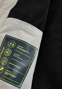 Tissu gris clair et noir avec une étiquette rectangulaire décrivant les caractéristiques imperméables et coupe-vent ainsi que les avantages des coutures scellées en texte jaune.