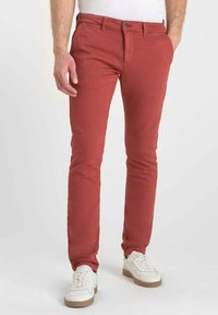 Man som bär slim-fit röda byxor och vita sneakers, stående med en hand i fickan mot en enfärgad bakgrund.