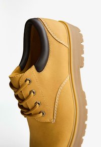 Pantof din suede galben, cu accente din piele maro. Dispune de șase orificii metalice, șireturi rotunde și o talpă robustă, texturată.