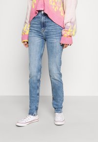 Ljust blå denimjeans med slim fit, som har hög midja och subtil blekning. Kombineras med en stickad tröja i rosa och gult mönster samt vita sneakers.