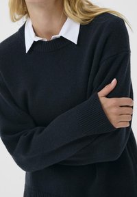 Navy knit sweater with a round neckline and ribbed cuffs. Layered over a white collared shirt, showcasing a textured finish.
