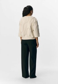 Veste beige avec des lignes texturées et en relief, manches trois-quarts, associée à un pantalon large noir. Design simple avec une coupe décontractée.