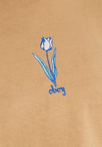 Beige tygtröja med en broderad tulpandesign i blått och rosa och texten "obey" nedanför i blå bokstäver.