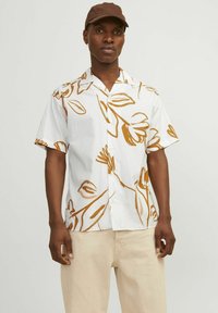 Camicetta a maniche corte di colore bianco con un motivo floreale marrone. Materiale in cotone, design con bottoni e vestibilità comoda. Abbinata a pantaloni beige chiaro.