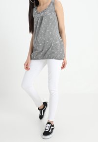 Kvinna som bär en ärmlös svart och vit randig topp, vita byxor och svarta sneakers, stående mot en vit bakgrund.