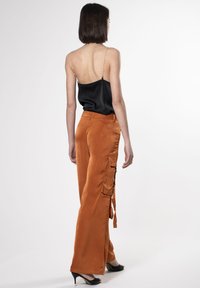 Schwarzes Satin-Camisole mit dünnen Trägern; orangefarbene weite Hosenträger mit Cargo-Taschen und seitlichen Bindebändern. Modell trägt schwarze High-Heels.