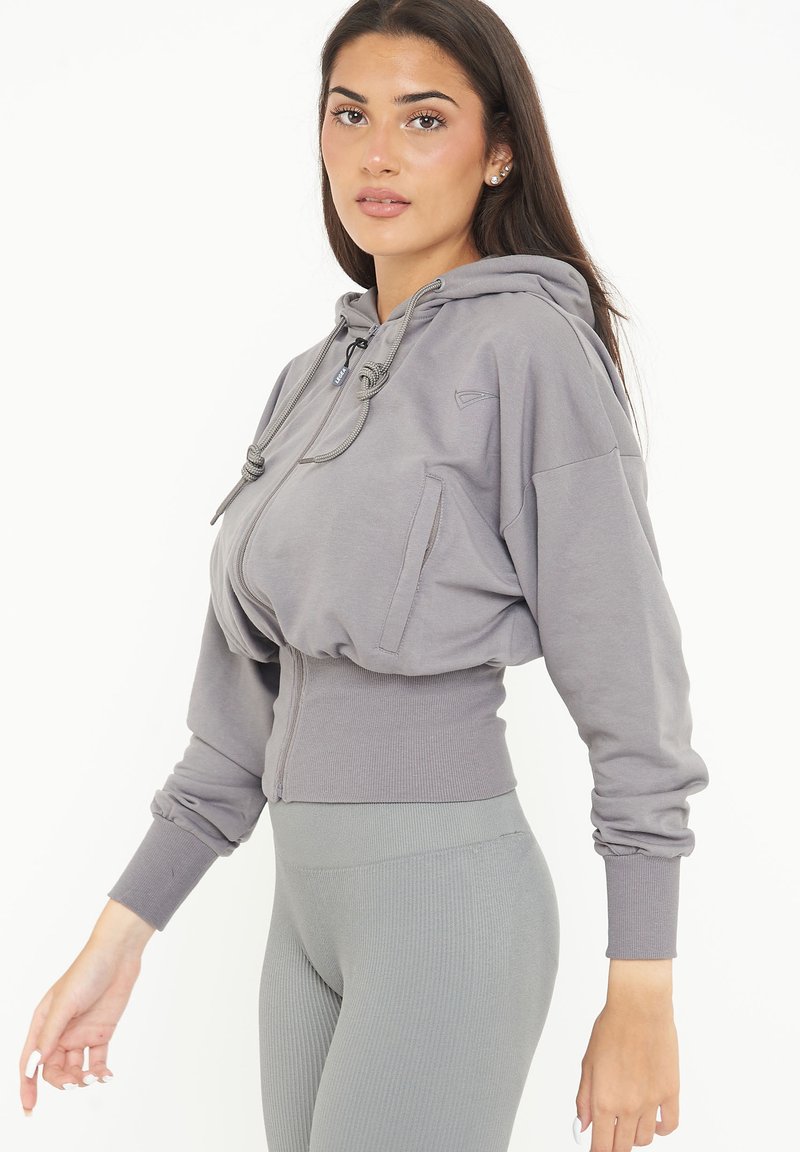 Hoodie grigio corto con lacci, tasca frontale e orlo a coste. Abbinato a leggings grigi a coste coordinati. Texture liscia, vestibilità rilassata.